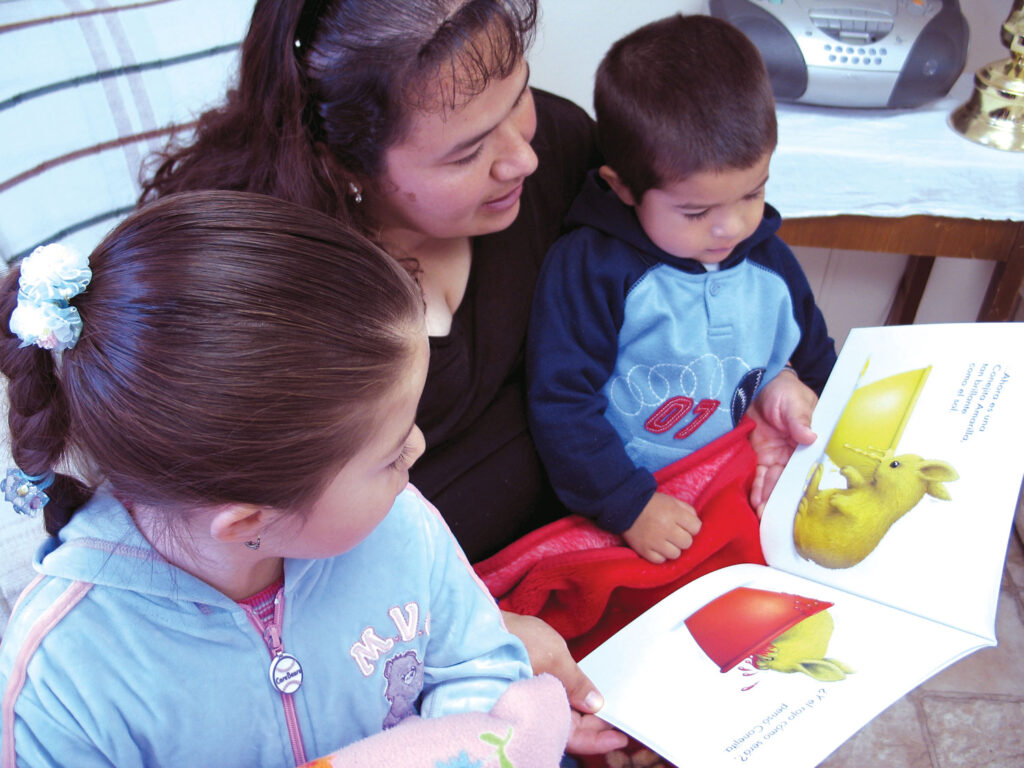 A woman reads a colorful book to two children sitting on her lap
